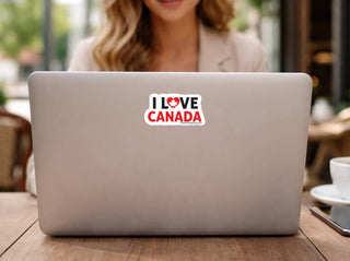 A white die-cut sticker with the text 'I LOVE CANADA' and a red heart, shown on the back of a silver laptop. The laptop is on a wooden table in a cafe setting with a woman blurred in the background.