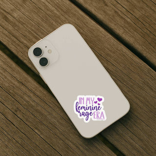 In My Feminine Rage Era feminist sticker with purple script applied to the back of a light-colored smartphone on a wooden surface.