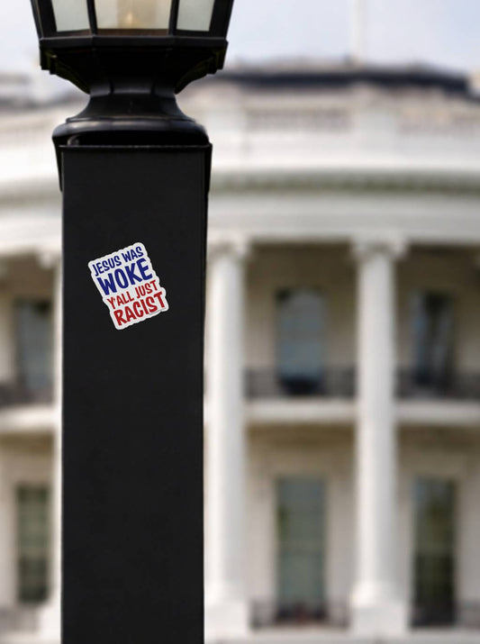 A "Jesus Was Woke" sticker placed on a black lamp post with the blurred North Portico of the White House in the background.