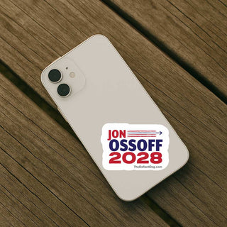 A white phone case with a red and blue 'Jon Ossoff 2028' campaign sticker, shown on a light beige smartphone resting on a dark brown wooden plank surface.