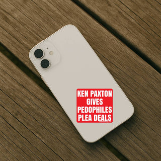 A white smartphone case shown from the back, placed on a wooden surface. The case has a red rectangular sticker with white text reading 'KEN PAXTON GIVES PEDOPHILES PLEA DEALS.
