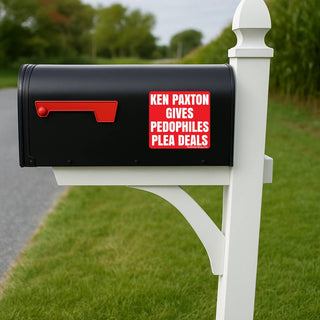 A black mailbox mounted on a white post outdoors, with a red rectangular sign on the front that reads “KEN PAXTON GIVES PEDOPHILES PLEA DEALS.” The background shows green grass, trees, and a blurred road.