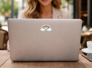 Love Always Wins rainbow sticker centered on the back of a neutral-toned laptop at an outdoor café setting. The design adds a bold yet cheerful pride statement to a clean tech aesthetic.