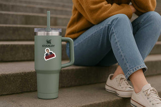 Person sitting on steps with a Stanley tumbler and a sticker on the cup