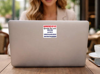 A silver laptop on a wooden table in a cafe setting, with a white rectangular sticker on the lid that reads 'MURDERED BY ICE Remember their names: RENEE GOOD, ALEX PRETTI, LORENZO SALGADO ARAUJO, JOAN SEBATIAN GUERRERO'. A blurred person is visible in the background.