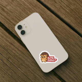 My Tummy Hurts and Im Mad at the Government hedgehog political humor sticker applied to the back of a light-colored smartphone on a wooden surface.