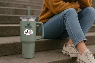 A sage green Stanley tumbler with a handle and straw sits on concrete steps next to a person sitting on steps wearing a mustard sweater, blue jeans, and white sneakers.