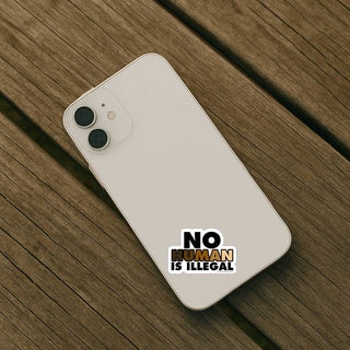 No Human Is Illegal sticker displayed on the back of a light-colored smartphone resting on a wooden surface. The compact design shows how the sticker fits cleanly on smaller personal items.