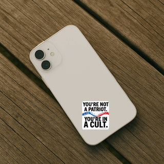 Youre Not a Patriot Youre in a Cult political statement sticker applied to the back of a light-colored smartphone on a wooden surface.