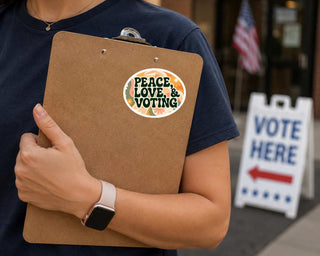 A person in a navy blue t-shirt holds a brown cardboard clipboard with a circular sticker that reads 'PEACE LOVE & VOTING' surrounded by autumn leaves. In the background, an American flag hangs on a building, and a white 'VOTE HERE' sign with a red arrow stands on the sidewalk.