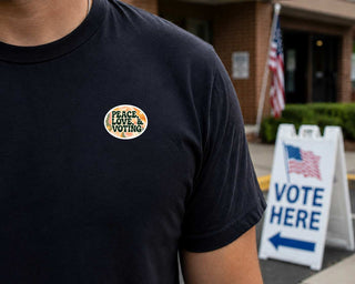 A close-up of a person wearing a black t-shirt with a circular 'PEACE LOVE VOTING' pin on the chest, standing outdoors near a 'VOTE HERE' sign and an American flag.