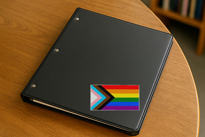 Progressive Gay Pride Flag' magnet or vinyl decal displayed on a black binder in a library. A bold, high-quality political sticker perfect for cars, laptops, fridges, or toolboxes. Great for activists, protestors, or anyone fed up with fascism.