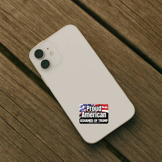 An overhead shot of an off-white iPhone lying on a weathered wooden surface. The "Proud American Ashamed of Trump" sticker is applied to the lower center of the phone's back.