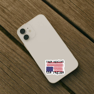 Rage Against the Regime American flag protest sticker applied to the back of a light-colored smartphone on a wooden surface.