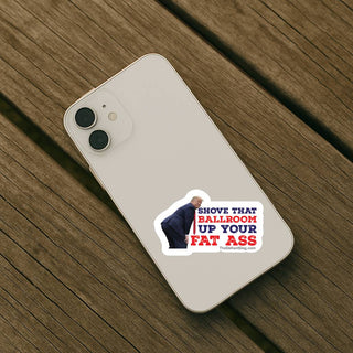Die-cut anti-Trump political sticker on the back of a light-colored smartphone resting on a wooden table, featuring Donald Trump and the Shove That Ballroom Up Your Fat Ass design.