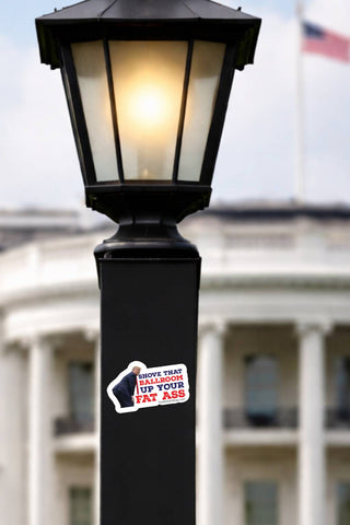 Anti-Trump ballroom protest sticker applied to a black outdoor lamp post with a government-style white building and American flag blurred in the background.