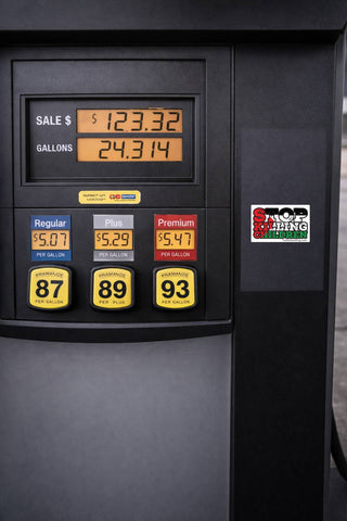 Close-up of a black gas pump display showing sale price, gallons, and three fuel options labeled Regular, Plus, and Premium with yellow price badges. A Stop  KILLING CHILDREN sticker is visible on the side.