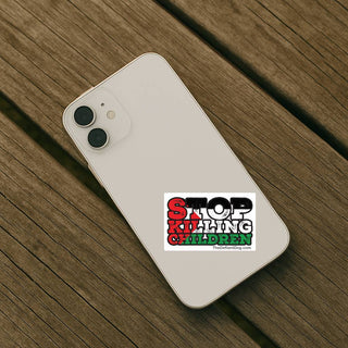A white smartphone lying on a weathered wooden plank surface, angled diagonally. On the back of the phone is a rectangular sticker with the text 'STOP KILLING CHILDREN' in red, black, and green lettering.