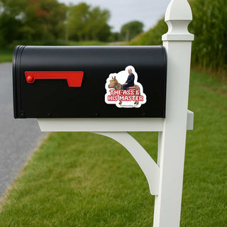 A black mailbox mounted on a white post outdoors, with a red flag and a die-cut sticker on the side showing a man and dog with the text 'THE ASS & HIS MASTER'.