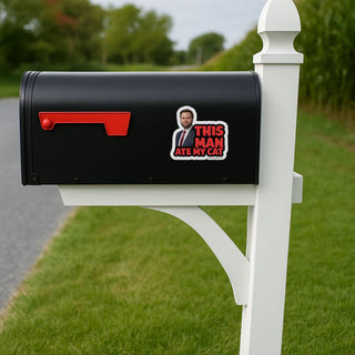 Black mailbox with a sticker on it, featuring a man and text, in a grassy area.