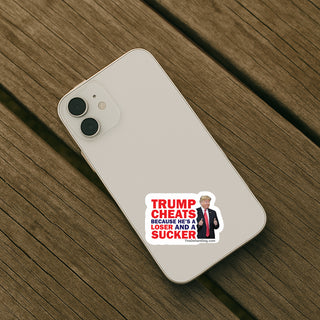 A white smartphone case lying on a wooden surface, printed with a political sticker design featuring the text 'TRUMP CHEATS BECAUSE HE'S A LOSER AND A SUCKER' in red and white, plus a small photo of Donald Trump.