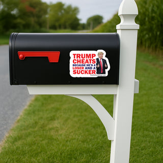 A black mailbox mounted on a white post outdoors, with a rectangular political sticker on the front reading 'TRUMP CHEATS BECAUSE HE'S A LOSER AND A SUCKER' and showing a portrait of Donald Trump.