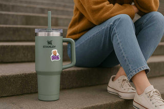 Person sitting on steps with a green Stanley tumbler and a sticker, wearing a mustard sweater and blue jeans.