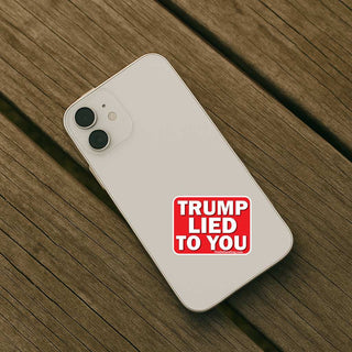 A white smartphone lying diagonally on a weathered wooden plank surface, featuring a red square sticker on the back with the text 'TRUMP LIED TO YOU' in white capital letters.