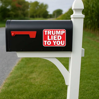 A black mailbox mounted on a white post outdoors, with a red flag and a red-and-white sticker that reads “TRUMP LIED TO YOU.” The background shows grass, trees, and a blurred road.