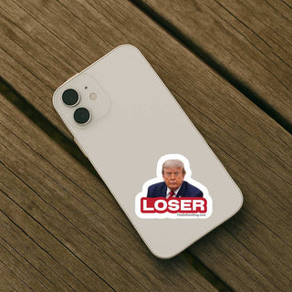 A white smartphone case shown from the back, placed on a brown wooden surface. The case has a printed sticker near the lower center featuring a portrait of Donald Trump above the word 'LOSER' in red and white, with a small website URL underneath.