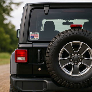 The rear corner of a black Jeep Wrangler. The "UNDER DISTRESS" upside-down flag decal is applied to the lower-left corner of the rear window, next to the spare tire. A red brake light is visible above the tire.