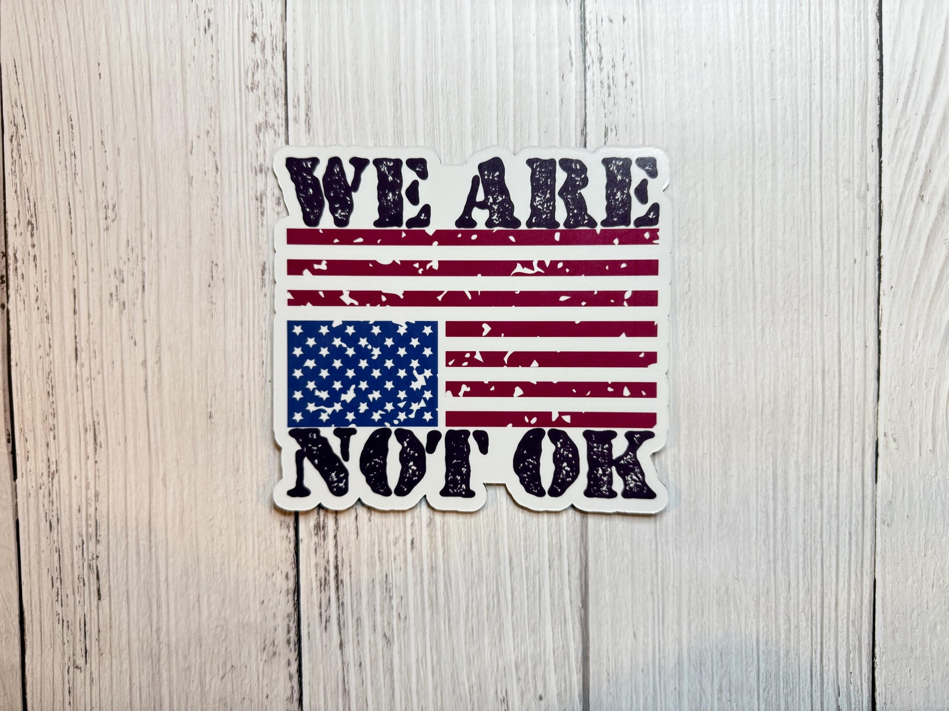 A vinyl decal or magnet featuring the phrase "We are not OK" in bold lettering, displayed alongside or over an upside-down American flag. The design symbolizes national distress and conveys a message of concern, protest, and social or political unrest.