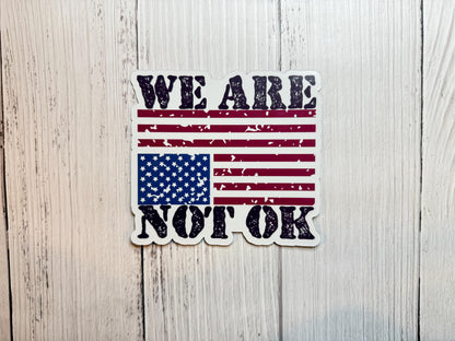A vinyl decal or magnet featuring the phrase "We are not OK" in bold lettering, displayed alongside or over an upside-down American flag. The design symbolizes national distress and conveys a message of concern, protest, and social or political unrest.
