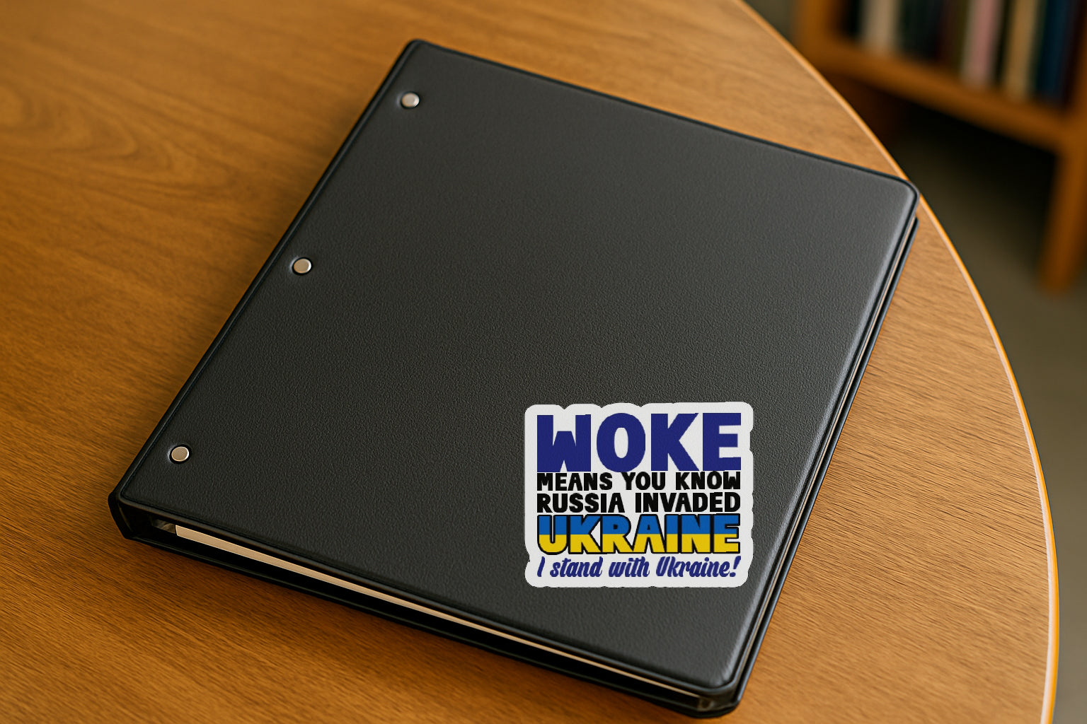 Woke Means You Know Russia Invaded Ukraine  I Stand with Ukraine' magnet or vinyl decal displayed on a black binder in a library. A bold, high-quality political sticker perfect for cars, laptops, fridges, or toolboxes. Great for activists, protestors, or anyone fed up with fascism.