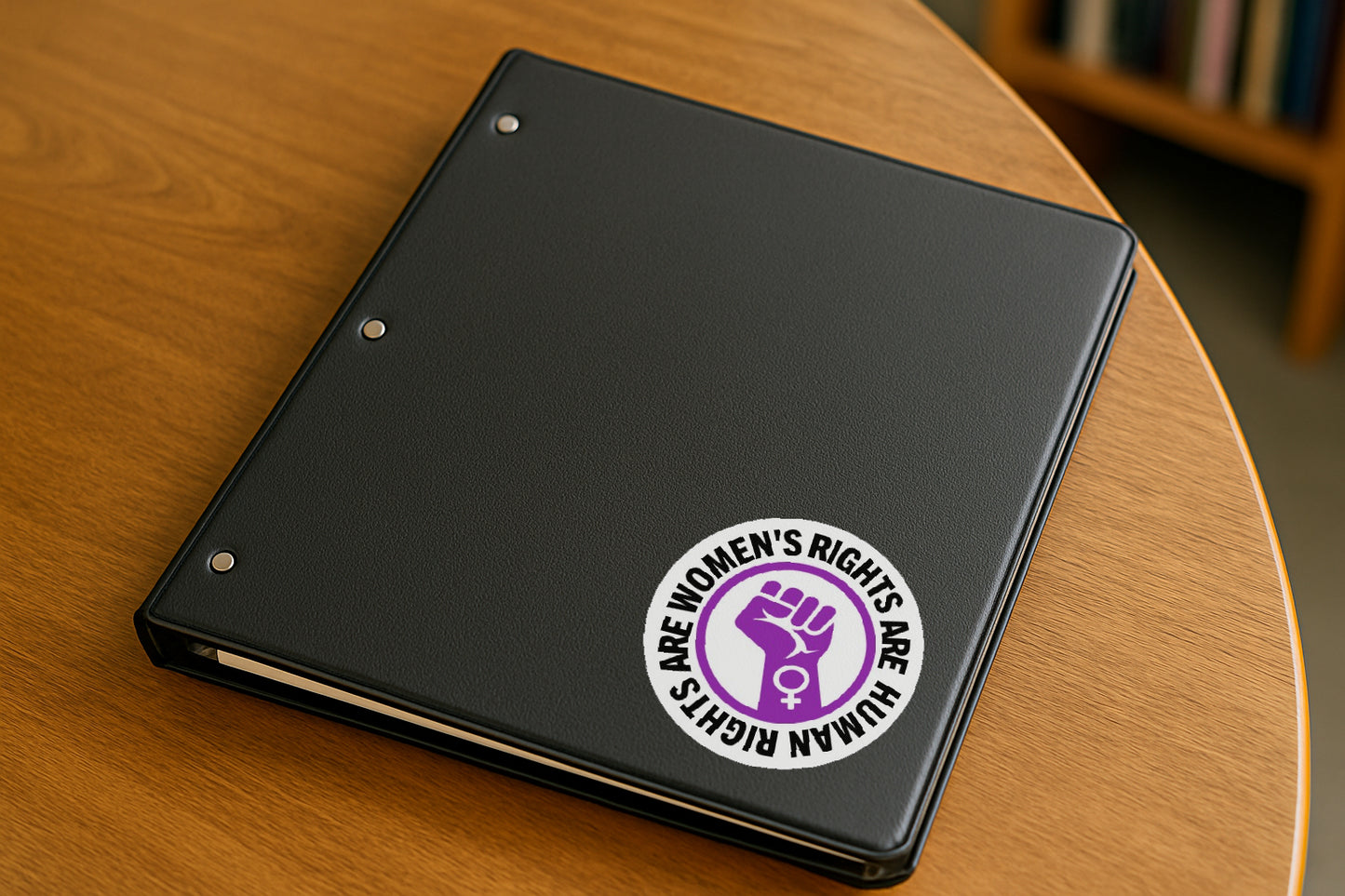Women's Rights Are Human Rights' magnet or vinyl decal displayed on a black binder in a library. A bold, high-quality political sticker perfect for cars, laptops, fridges, or toolboxes. Great for activists, protestors, or anyone fed up with fascism.