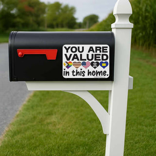 Black mailbox with a 'You are valued in this home' sticker on a white post, outdoors.