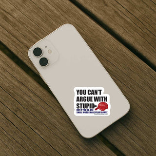 A white smartphone case with a humorous sticker on it, resting on a dark brown wooden plank surface. The sticker features the text 'YOU CAN'T ARGUE WITH STUPID' and a red cap illustration with 'MAGA' on it. Smaller blue text below says 'BUT IF YOU DO, USE SMALL WORDS AND SPEAK SLOWLY.