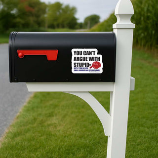 A black mailbox mounted on a white post outdoors, with a red flag and a white sticker that reads 'YOU CAN'T ARGUE WITH STUPID' and a red cap illustration with 'MAGA' on it. Smaller blue text below says 'BUT IF YOU DO, USE SMALL WORDS AND SPEAK SLOWLY. The background shows grass, trees, and a road.