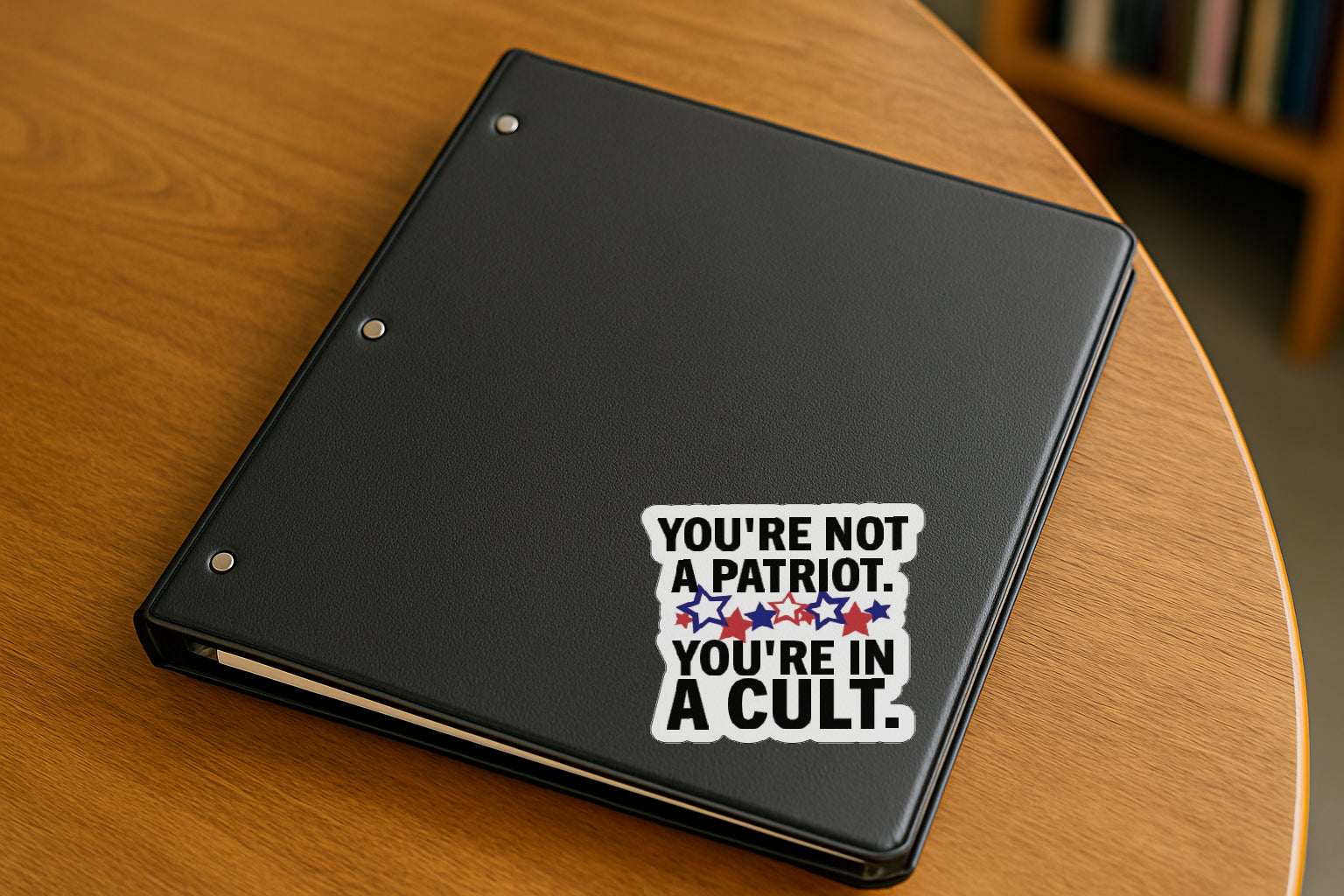 You're Not a Patriot  You're in a Cult' magnet or vinyl decal displayed on a black binder in a library. A bold, high-quality political sticker perfect for cars, laptops, fridges, or toolboxes. Great for activists, protestors, or anyone fed up with fascism.